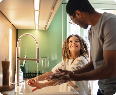 Vīrietis pie virtuves izlietnes rāda bērnam, kā pareizi mazgāt rokas, kas ir daļa no veselīgas personīgās higiēnas.